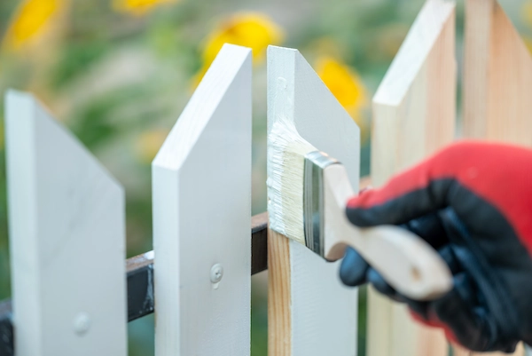 Painter wearing red and black gloves applying white paint to a wooden picket fence with a brush