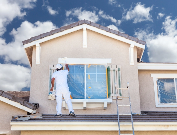House painter in white clothing standing on a rooftop painting the exterior trim around a window with plastic sheeting taped over the glass