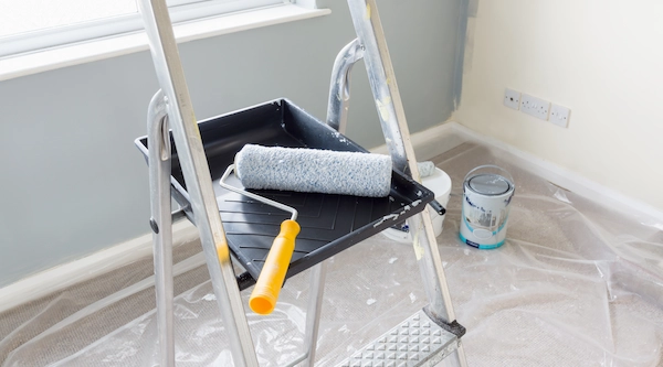 Paint roller and tray on a stepladder with a can of gray paint and protective plastic sheeting on the floor during an interior room painting project