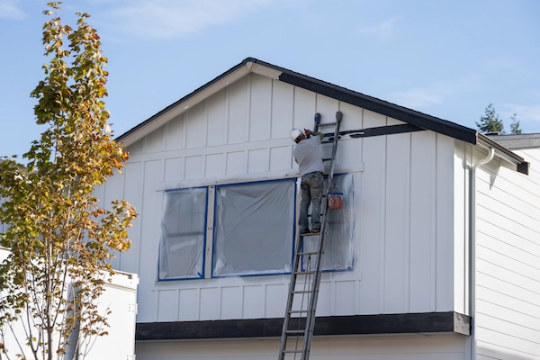 Painter on a tall ladder painting the upper exterior of a white board-and-batten house with windows covered in plastic sheeting